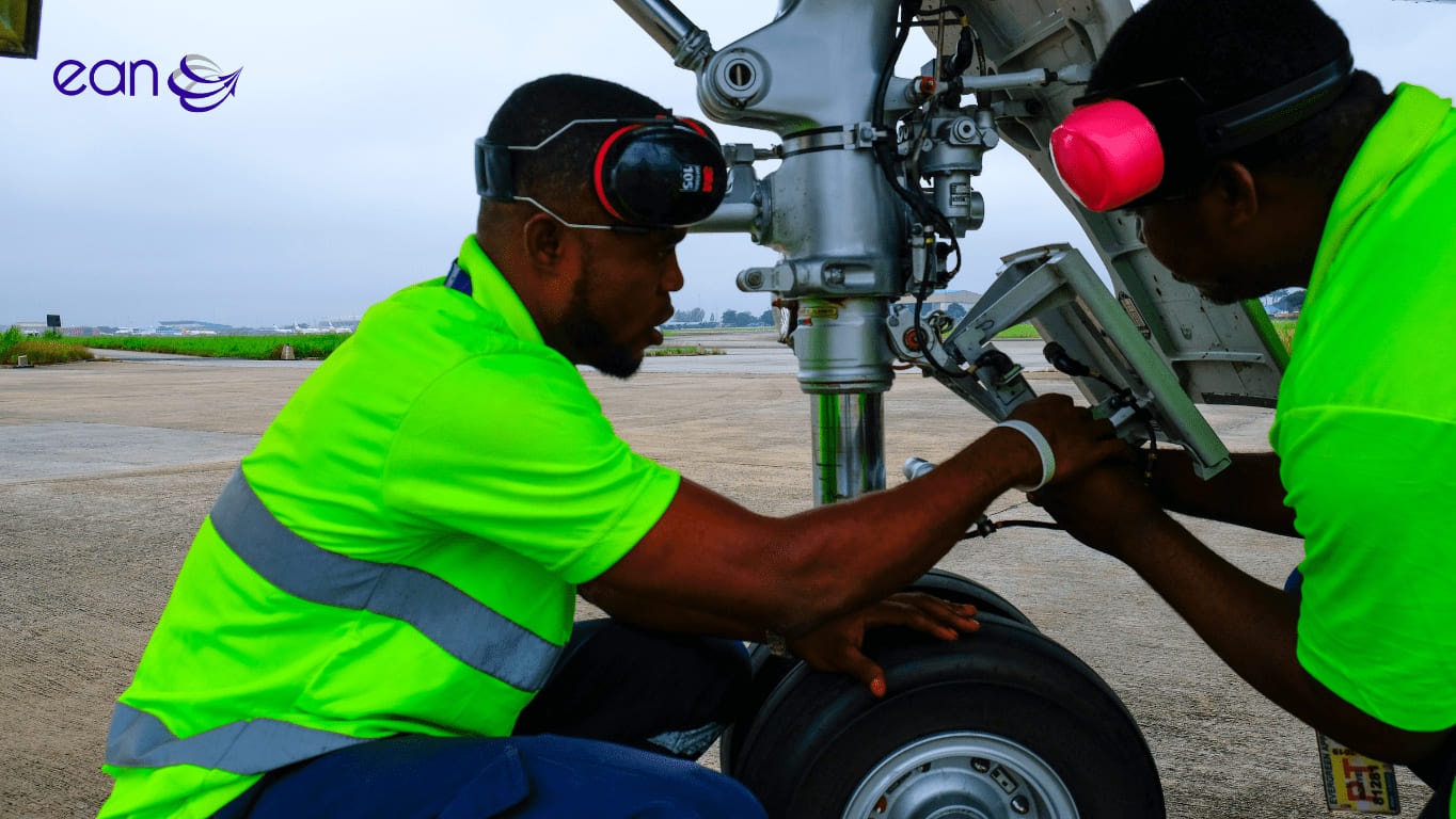 Two EAN engineers in high-visibility shirts and ear defenders working on an aircraft’s nose landing gear on the ramp.