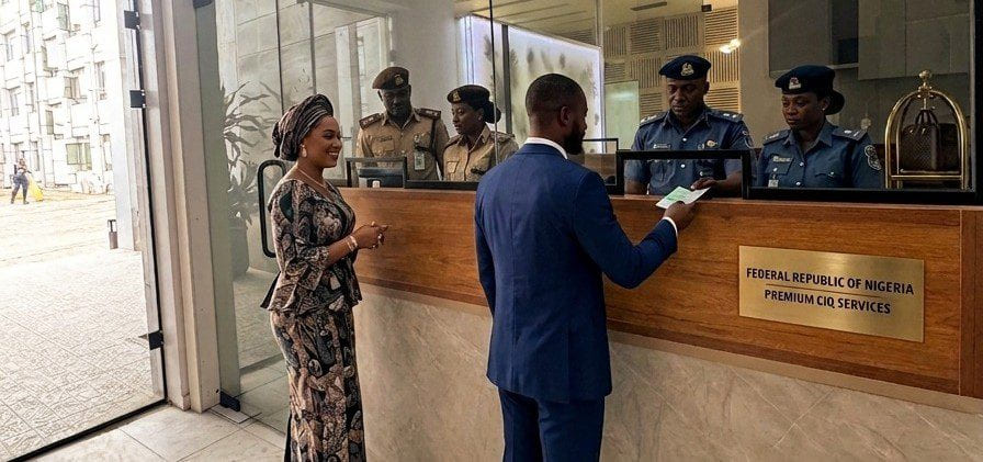 A traveller handing a passport across a counter signed “Federal Republic of Nigeria — Premium CIQ Services”, staffed by uniformed immigration and customs officers.